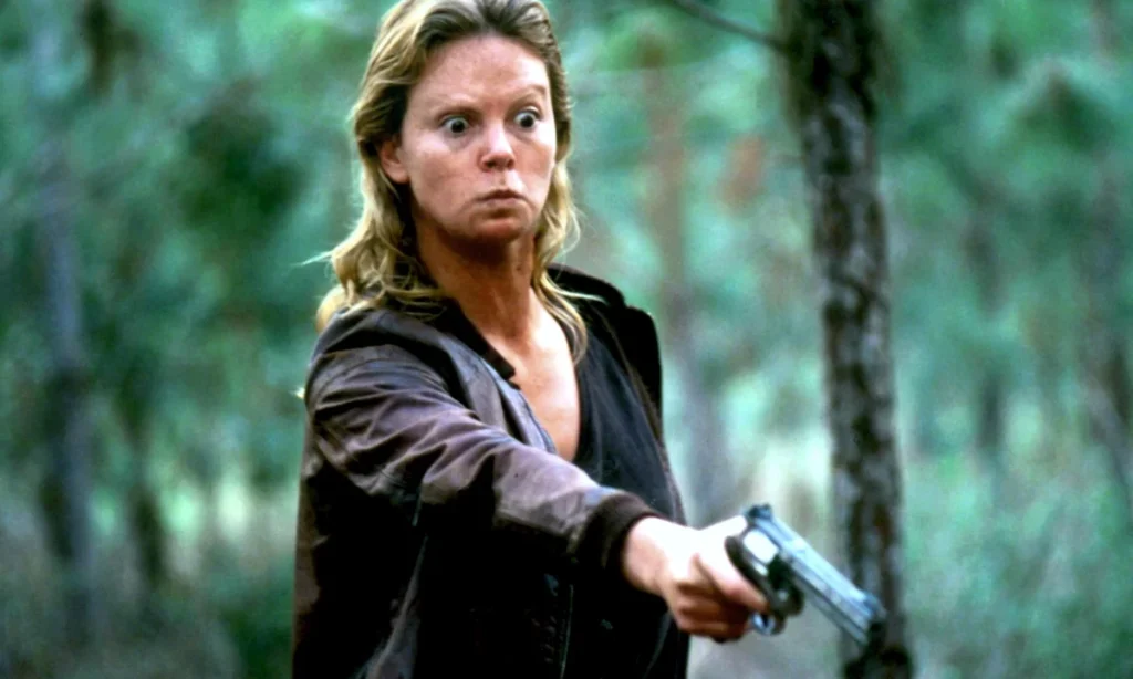 Charlize Theron as Aileen Wuornos, with messy blonde hair and a surprised expression, pointing a revolver in a forest setting in "Monster."