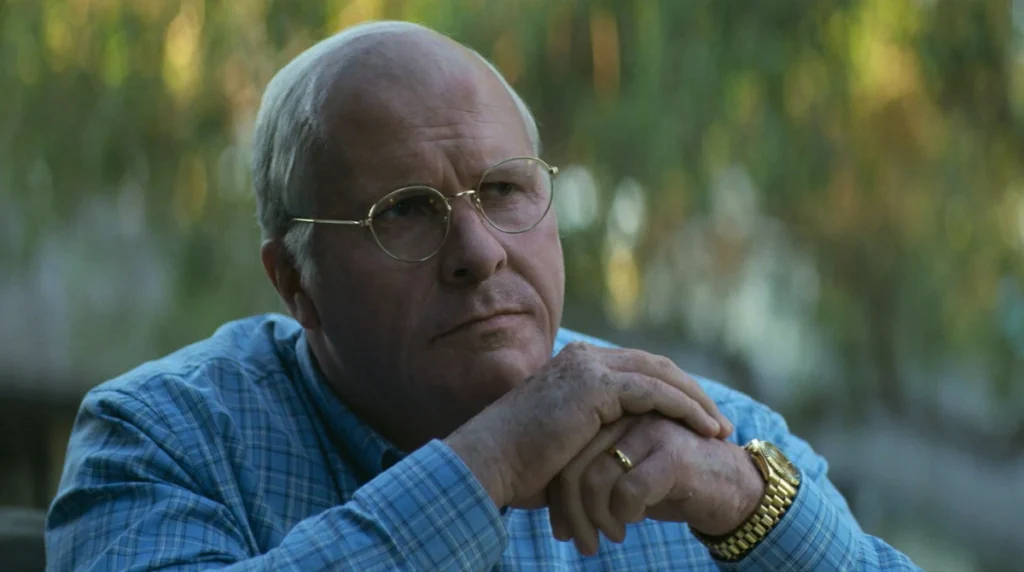 Christian Bale as Dick Cheney, wearing round glasses and a blue plaid shirt, with his hands clasped under his chin, looking contemplative in "Vice."