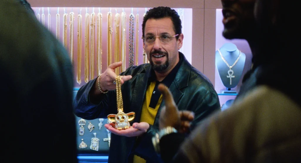 Adam Sandler as Howard Ratner, wearing glasses and a goatee, holding up a large gold Furbie pendant for a customer in his jewelry store in "Uncut Gems."