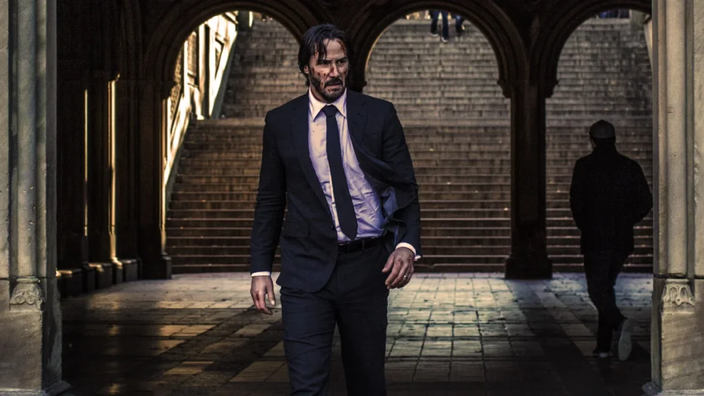 John Wick (Keanu Reeves) walking determinedly through a shadowy archway, looking battle-worn.