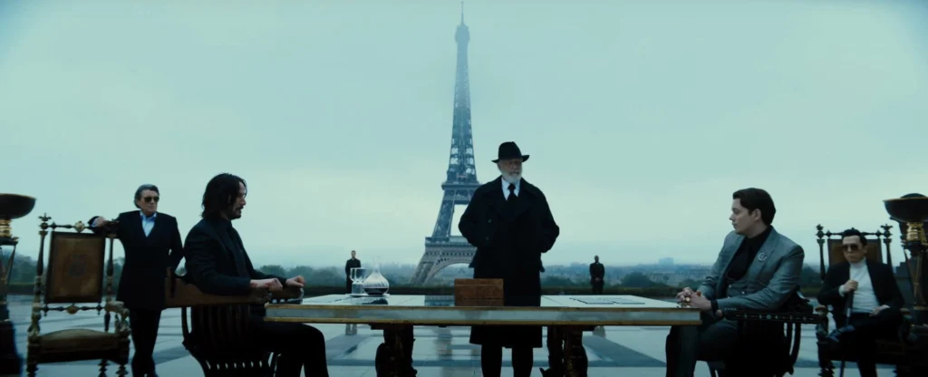 ohn Wick, Winston, the Marquis, and other characters seated at a table outdoors in Paris, with the Eiffel Tower visible in the distance.