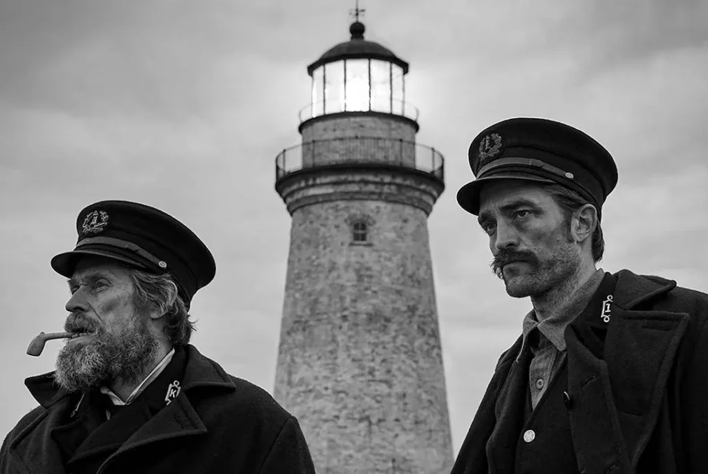 Willem Dafoe and Robert Pattinson as lighthouse keepers, standing in uniform in front of a lighthouse in "The Lighthouse."
