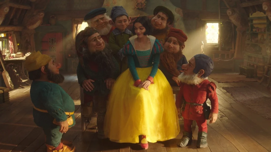 Rachel Zegler as Snow White in a yellow dress and blue top, standing with six diverse magical creatures in a rustic cottage setting.
