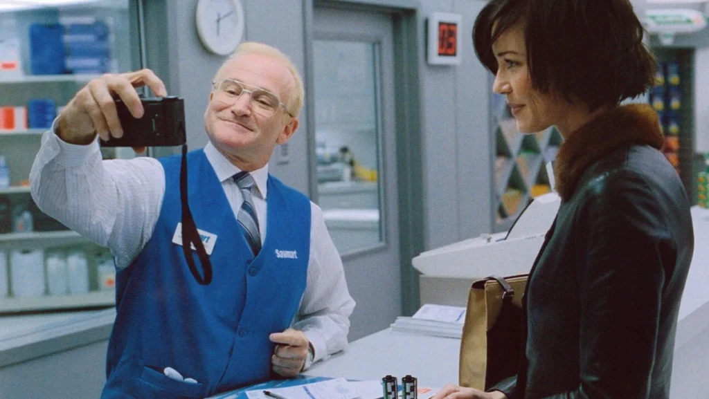Robin Williams as Sy Parrish, a smiling photo technician in a blue vest, holding up a camera to a female customer in "One Hour Photo."