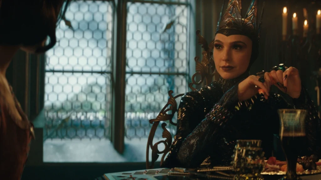 Gal Gadot as the Evil Queen, wearing a dark, ornate gown and crown, sitting at a table with a sly expression.