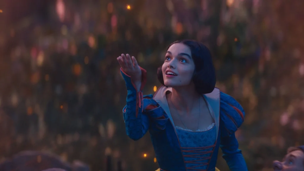 Rachel Zegler as Snow White looking upwards with a look of wonder, surrounded by glowing fairy-like lights in a magical forest.